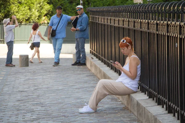 Uma turista menina senta-se perto da cerca e olha para o smartphone e outros turistas passam . — Fotografia de Stock