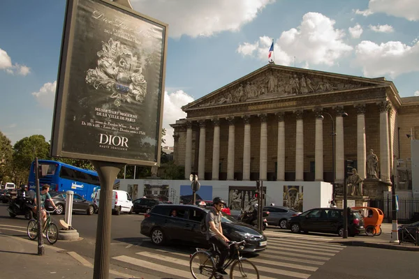 Ciclistas y coches recorren la Asamblea Nacional con el telón de fondo de un cartel de Dior . — Foto de Stock