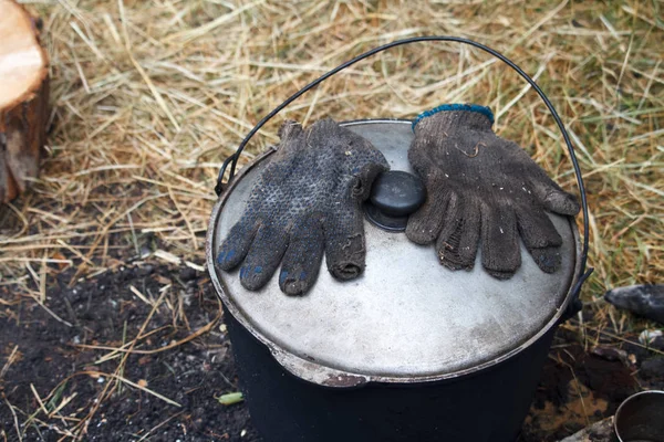 Gants en suie sont sur le couvercle d'un pot avec de la nourriture prête à l'emploi près du feu — Photo