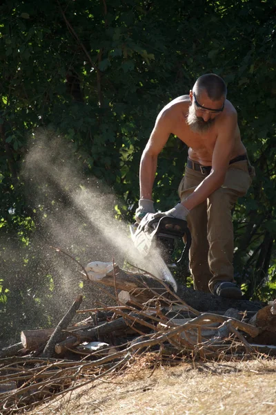 Barefoot man with a beard cuts firewood for the fire by chainsaw in the morning sun — Stock Photo, Image