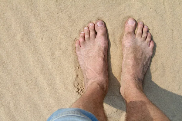 Close-up of male feet and legs in denim shorts are standing on the white sand on the beach or in the desert — Stock Photo, Image