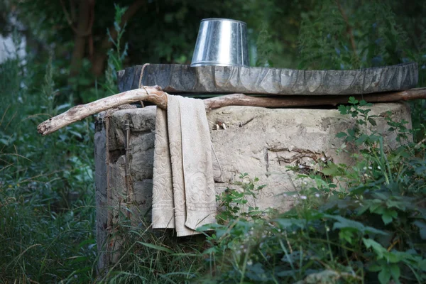 Un seau vide brillant est retourné à l'envers sur le couvercle d'un puits, une vieille serviette est accrochée à côté sur un fond de jardin vert flou — Photo