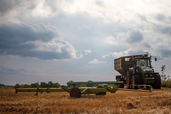 Sebuah traktor untuk mengumpulkan gandum dari pengumpul gabungan berdiri di ladang gandum yang dipangkas melawan langit yang berawan biru — Stok Foto