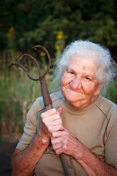 Nahaufnahme Porträt einer alten Frau mit grauen Haaren, die eine rostige Mistgabel oder einen Chopper in den Händen hält, Gesicht in tiefen Falten, selektiver Fokus — Stockfoto