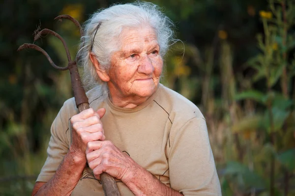 Close-up retrato de uma mulher idosa com cabelos grisalhos segurando um garfo enferrujado ou helicóptero em suas mãos, rosto em rugas profundas, foco seletivo — Fotografia de Stock