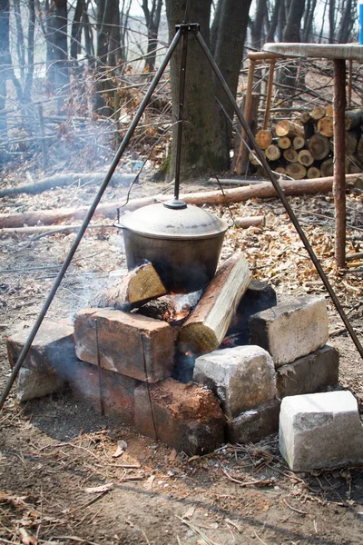 The concept of camping kitchen, on a tripod over a fire there is a pot in which food is prepared on the background of a pile of dry branches