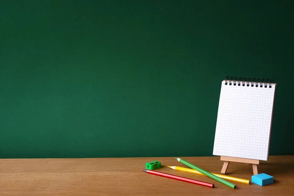 Okul konseptine geri dönelim, minyatür şelnet üzerine açık defter ve temiz yeşil tebeşir tahtasının zemininde ahşap yüzeyde birkaç renkli kalem, seçici odak, kopyalama alanı