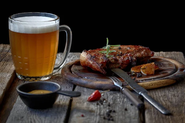 Büyük bir kızarmış biftek, bir bardak bira, hardal ve çatal bıçak takımı ile eski bir ahşap masa, Oktoberfest ve St Patrick Günü kavramı ile natürmort