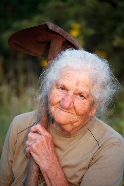 Close-up portret van een oude vrouw met grijs haar glimlachend en kijkend naar de camera, vasthouden van een roestige schop in haar handen, gezicht in diepe rimpels, selectieve focus — Stockfoto