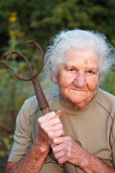 Close-up retrato de uma mulher idosa com cabelos grisalhos segurando um garfo enferrujado ou helicóptero em suas mãos, rosto em rugas profundas, foco seletivo — Fotografia de Stock