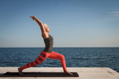 Kısa saç kesimi uygulamaları ile yetişkin sarışın kadın deniz ve mavi gökyüzü arka plan karşı iskelede yoga