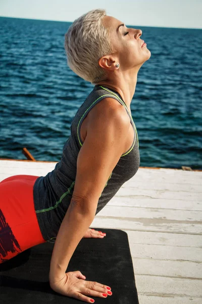 Potret close-up dari wanita pirang dewasa dengan potongan rambut pendek berlatih yoga di dermaga dengan latar belakang laut dan langit biru — Stok Foto