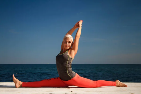 Wanita pirang dewasa dengan potongan rambut pendek berlatih yoga di dermaga dengan latar belakang laut dan langit biru — Stok Foto