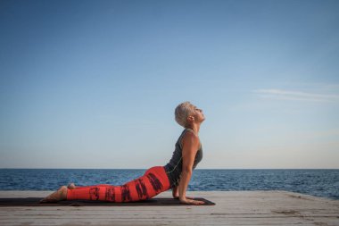 Kısa saç kesimi uygulamaları ile yetişkin sarışın kadın deniz ve mavi gökyüzü arka plan karşı iskelede yoga