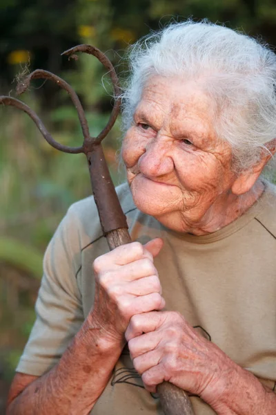 Nahaufnahme Porträt einer alten Frau mit grauen Haaren, die eine rostige Mistgabel oder einen Chopper in den Händen hält, Gesicht in tiefen Falten, selektiver Fokus — Stockfoto