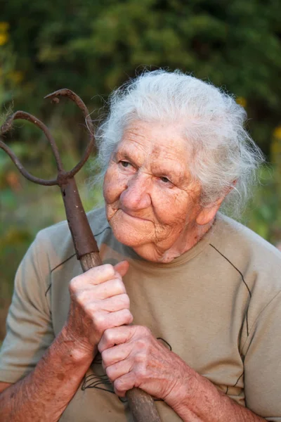 Close-up retrato de uma mulher idosa com cabelos grisalhos segurando um garfo enferrujado ou helicóptero em suas mãos, rosto em rugas profundas, foco seletivo — Fotografia de Stock