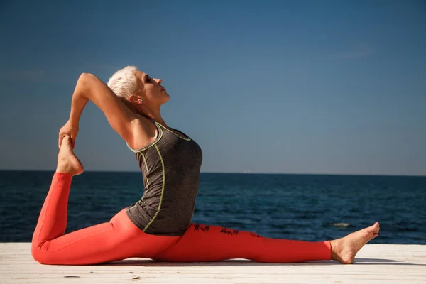 Wanita pirang dewasa dengan potongan rambut pendek berlatih yoga di dermaga dengan latar belakang laut dan langit biru — Stok Foto