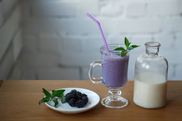 Batido de mora de cerca en una taza de vidrio, una botella de leche y un platillo con bayas frescas y una ramita de menta, enfoque selectivo — Foto de Stock