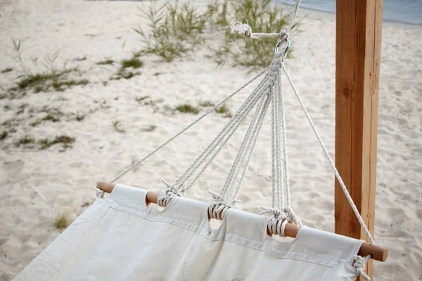 Крупный план белого гамака из натуральной ткани на деревянном основании, висящем на канатах против песчаного пляжа, концепция естественного расслабления Лицензионные Стоковые Изображения