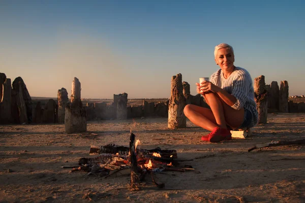 夕日の海のビーチでカップで火のそばに座っているセーターの大人の中年のブロンドの女性、選択的な焦点 — ストック写真