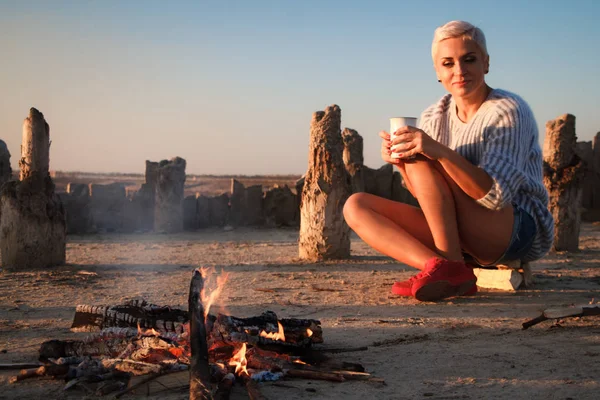 夕日の海のビーチでカップで火のそばに座っているセーターの大人の中年のブロンドの女性、選択的な焦点 — ストック写真