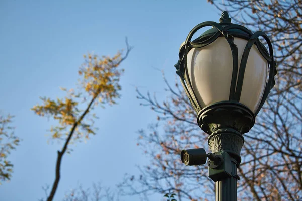 Vintage lantern with a surveillance camera in a public park in autumn in front of a blue sky, selective focus — Stock Photo, Image