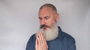 Olgun sakallı adam ellerini sıktı ve dua etti, dudaklarını oynattı, tevazu ve huzur, gri arka plan.