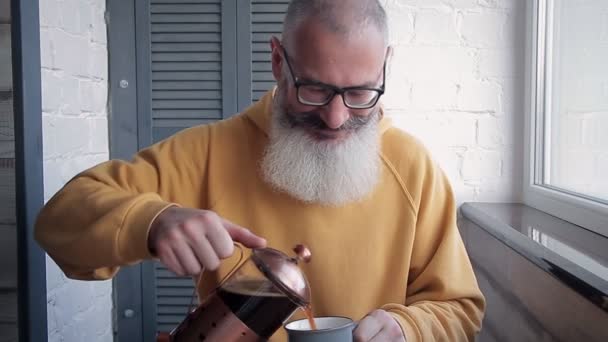 Felice uomo barbuto maturo versa troppo caffè in tazza e tende bere alla macchina fotografica. Sfondo cucina offuscata . — Video Stock
