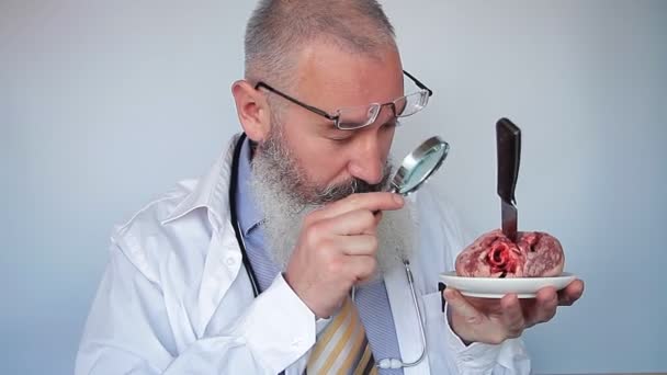 Зрілий бородатий чоловік лікар дивиться через збільшувальне скло в серці з ножем на тарілці. Дивна концепція лікаря. Концепція хірургії серця. Сірий фон . — стокове відео