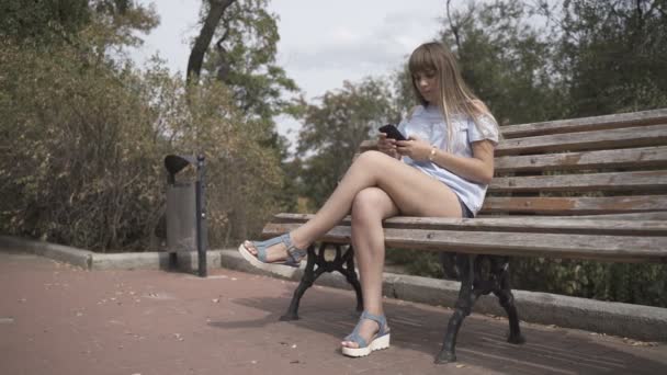 Девушка с телефоном сидит на скамейке — стоковое видео