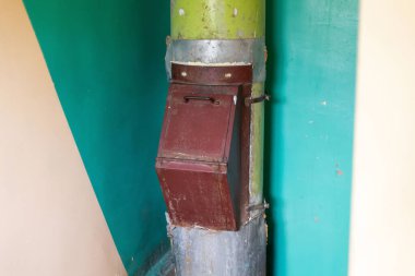 Bozulmakta olan bir binada paslanmış bir çöp kutusu yeşil metal bir sütuna yapıştırılır. Çevredeki duvarlar yıpranma belirtileri gösteriyor, ihmal edilen koşulları vurguluyor..