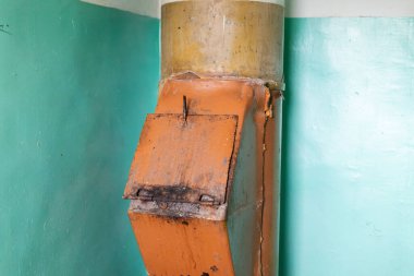 İhmal edilmiş bir çöp bacası bakımsız bir binada duruyor, paslı dış ve soluk turuncu rengini soyulmuş yeşil duvarlara gösteriyor, yıpranmış ve terk edilmiş yıllarını gösteriyor..
