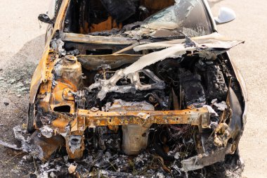 Yanmış bir araba otoparkta duruyor, geniş çaplı yangın hasarı var. Yanmış metal ve döküntüler, motor ve iç kısımların tahrip edildiğini gösteriyor. Turuncu boya kalıntıları da görünür..