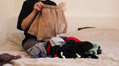 Bir kadın giysilerini yatağında düzgünce katlayarak düzenler. Konforlu yatak odası bu günlük ev işleri için öğleden sonraları rahat bir ortam sağlıyor..