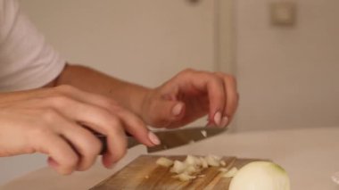 Sıcak bir mutfakta, bir kadın bir soğanı ustalıkla dilimler. Sahne, ellerinin ve doğrama tahtasının detaylarını yakalıyor. Yemek hazırlama sanatını vurguluyor..