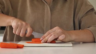 Sıcak bir mutfakta, bir kadın kesme tahtasındaki parlak turuncu havuçları ustalıkla dilimler. Taze malzemeler hazırlamaya odaklanması yemek pişirme sürecine hayat veriyor. Yetenek ve kendini adamışlık gösterisi..