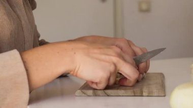 Bir kadın parlak bir mutfakta ahşap bir kesme tahtasıyla taze kereviz keser. Lezzetli bir yemek için gerekli malzemeleri hazırlarken ellere ve hassas hareketlere odaklanır..
