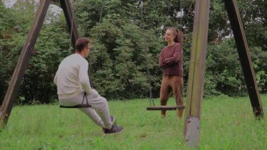 Bir çift yeşil bir parkta birlikte kaliteli zaman geçiriyorlar. Partnerlerden biri salıncakta şakacı bir şekilde otururken diğeri yakınlarda duruyor ve sohbet ediyor. Atmosfer rahat ve neşeli..