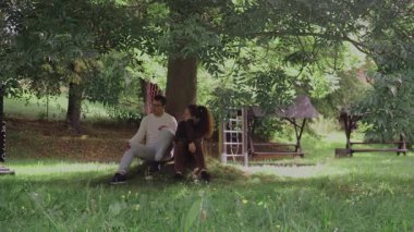 İki insan rahat bir anı paylaşıyor. Sakin bir parktaki büyük bir ağacın altında çimlerin üzerinde yan yana oturuyorlar. Kahkahaları ve konuşmaları doğanın güçlü bir bağını ve zevkini yansıtıyor.