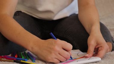 Bir kadın evde yaratıcı bir aktiviteye girişir, mum kalemlerle canlı ve soyut şekiller çizer. Kağıdı boyarken karmaşık tasarımlara odaklanıyor..