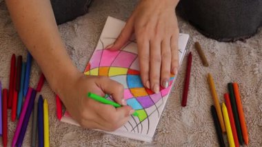 Bir kadın evinde rahat bir şekilde otururken balmumu boya kalemleri kullanarak canlı ve soyut şekiller çiziyor. Renkli çizgilere ve elleriyle yarattığı desenlere odaklanıyor..