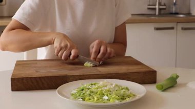 Parlak bir mutfakta, bir kadın keskin bir bıçakla taze pırasa kesmeye odaklanır. İnce kesilmiş yeşil ve beyaz parçalar, lezzetli bir yemek hazırlığı için bir kaseye konur..
