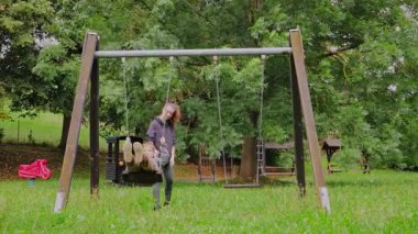 Genç bir anne, çocuğuyla hayat dolu bir oyun parkında salıncakta sallanıyor. Kahkaha ve eğlenceyi paylaşıyorlar. Yeşillik ve oyun malzemeleri arasında.