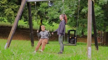 Huzurlu bir oyun alanında, genç bir anne çocuğuyla neşeyle sallanırken etkileşime girer. Bereketli yeşillikler bölgeyi kaplıyor ve eğlenceleri için mükemmel bir zemin oluşturuyor..