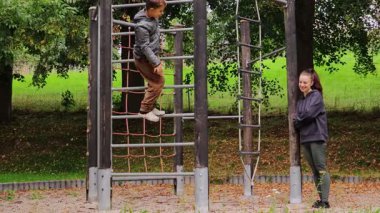 Çocuk parkında tırmanan genç bir anne çocuğuna destek olur. Sahnede yemyeşil ağaçlar ve güneşli bir öğleden sonra neşeli bir atmosfer var..