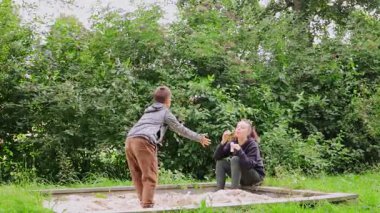 Genç bir anne ve çocuğu, yemyeşil yeşilliklerle çevrili oyun alanında harika vakit geçiriyorlar. Kum havuzunda oynuyorlar. Temiz hava ve güneş ışığının tadını çıkarıyorlar..