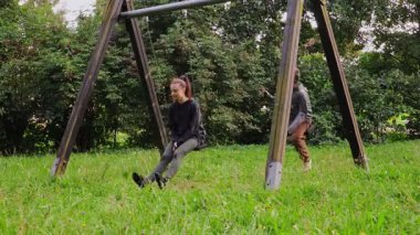 Huzurlu bir çocuk parkında, genç bir anne salıncakta sallanan bir çocukla neşeyle etkileşime geçer, yeşillikler arasında gülücükler saçar ve gülümser. Sıcaklık ve bağlantıyla dolu mükemmel bir gezinti..