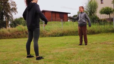 Arka bahçede, genç bir kadın çocuğuna çeşitli spor egzersizleri yaptırıyor, fitness ve bağ kurmayı teşvik ediyor. Kurulumda sıcak bir atmosfer oluşturan çimenlik bir alan ve birkaç ağaç yer alıyor..