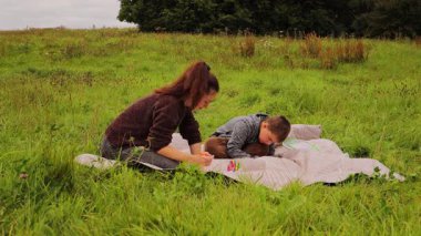 Genç bir kadın ve çocuğu yemyeşil çimlere serilmiş bir battaniyeye yaratıcı bir şekilde çizim yapıyorlar. Açık havada keyifli bir anı paylaşıyorlar, hayal gücünü arttırıyorlar..