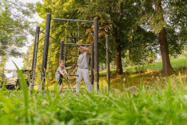 Bir baba ve çocuğu oyun alanında birlikte eğlenceli aktiviteler yaparak keyifli zaman geçiriyorlar. Canlı yeşil ağaçlar ve güneşli hava onların neşeli deneyimini güçlendirir..